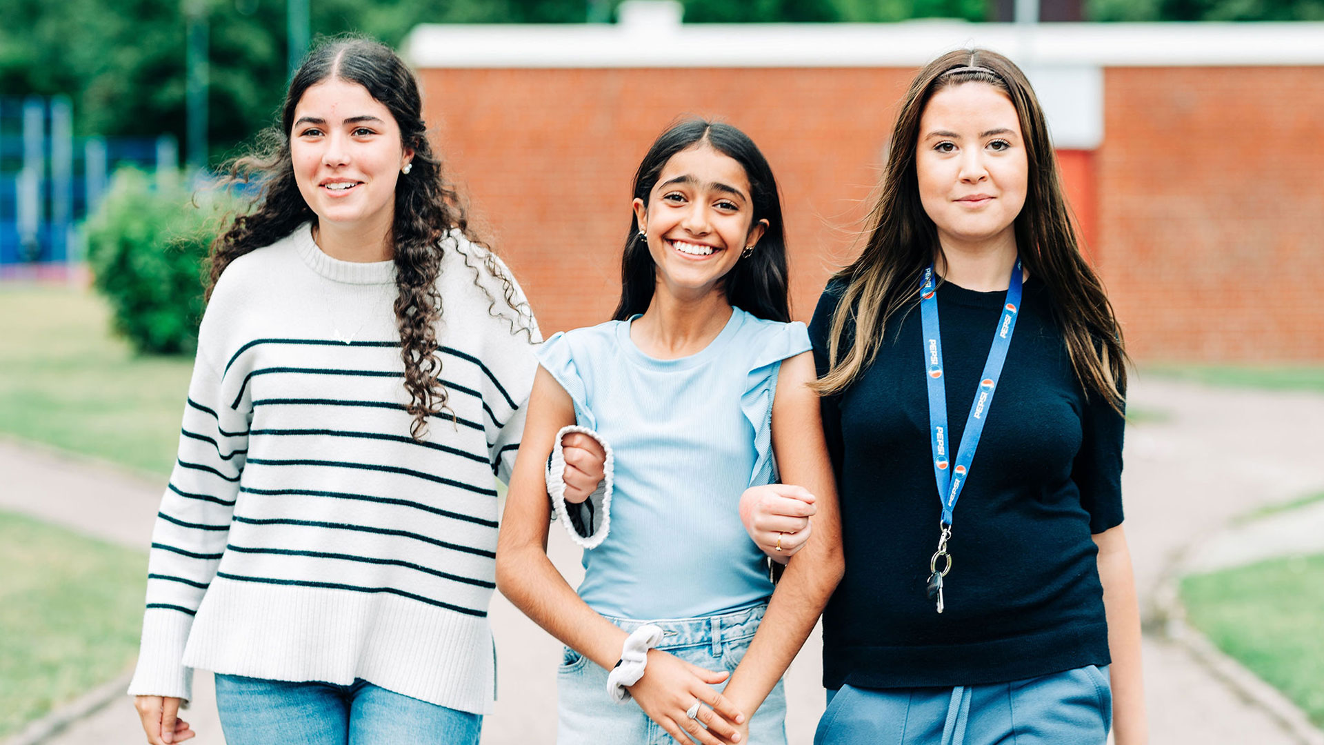 Tre unga kvinnor går tillsammans utomhus, arm i arm. De ler och verkar glada. I bakgrunden syns grönska och en tegelbyggnad.