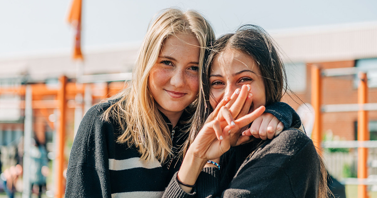 Två glada tjejer som står nära varandra utomhus. Den ena ler mot kameran, och den andra gör en rolig gest med handen. I bakgrunden syns en skolgård eller lekplats med orange detaljer.