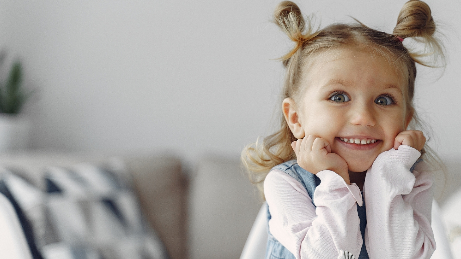 Leende flicka med uppsatt hår och ljusa kläder lutar sig glatt mot sina händer. Bakgrunden är ljus och suddig med en soffa och en kudde i bakgrunden.