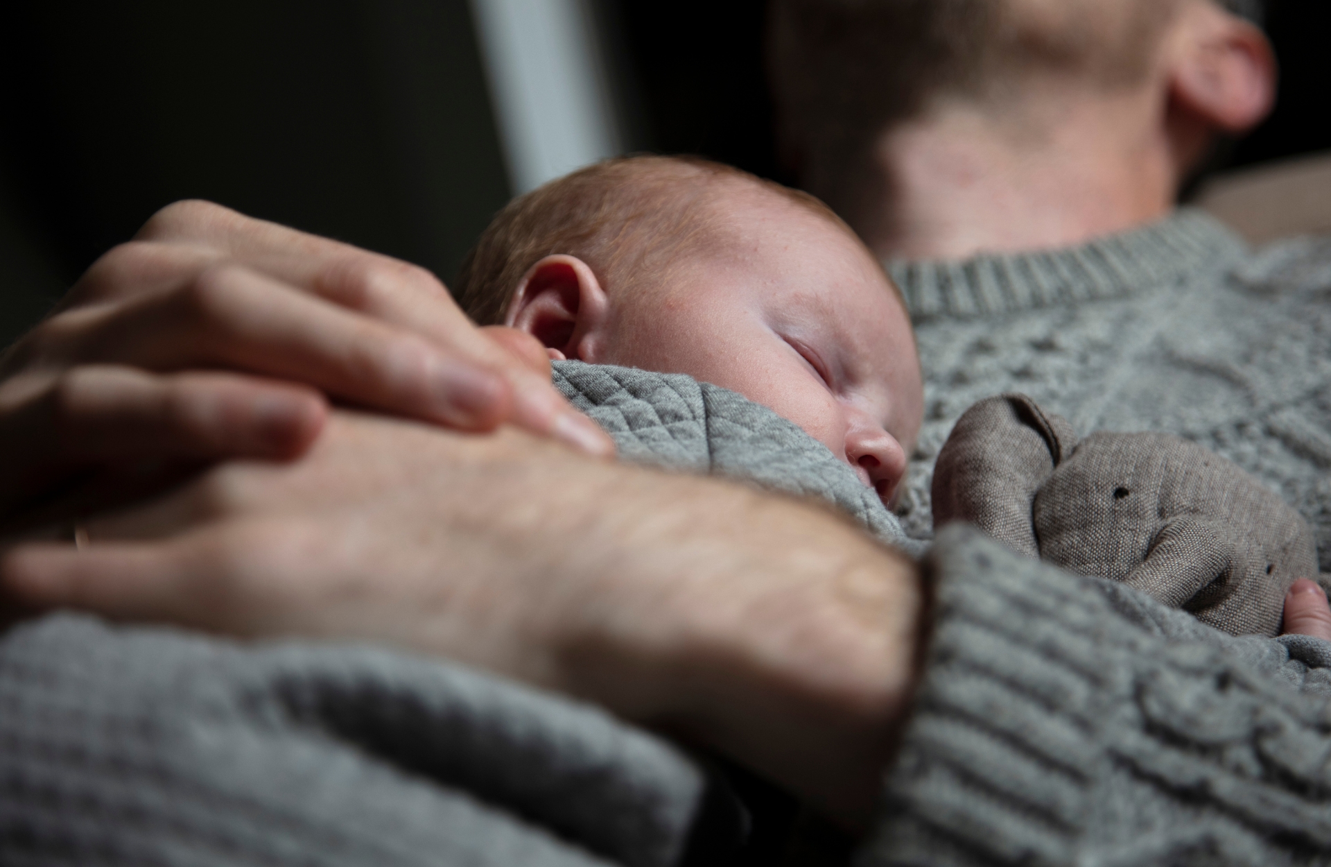 bebis-sover-tryggt-hos-pappa - 1920 x 1250 px En nyfödd bebis sover tryggt i famnen på en förälder, båda klädda i stickade grå tröjor. Närhet, trygghet och stillhet i hemmiljö.